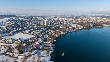 Fototapeta premium Aerial drone view along the shore of Hünenberg See shows snow-covered homes and residential blocks beside the deep blue Lake Zug under a clear, sunny winter sky.
