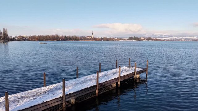 A drone glides slowly above a snow-covered wooden jetty along the western shore of Lake Zug in winter, with the church tower of Cham visible in the distance, creating a calm, meditative scene.