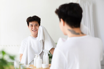 Fototapeta premium Morning procedures, male hygiene, beauty and skin care at home. Cheerful young asian guy wipes face with towel after washing and looks at reflection in mirror in white bathroom interior, free space