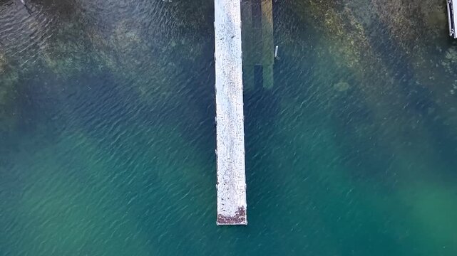 A snow-covered wooden jetty seen from above at Lake Zug, Switzerland, contrasting with turquoise-blue water and capturing a calm, minimalist winter atmosphere.