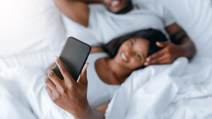 A couple is in bed, relaxed and smiling. The woman holds a smartphone and looks at it. The man lies...