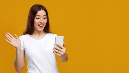 Excited young lady waving at mobile phone screen and smiling, having video conference with her girlfriends, happy to talk and to see friends, studio background, panorama with copy space