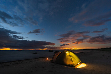 Illuminated camping tent on a sandy Baltic Sea beach in Estonia at night under a starry sky with sunset glow.