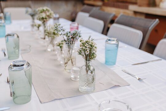 Close-up of small glass vases with wildflowers and wax flowers on a dining table. Rustic table decoration with colored water glasses
