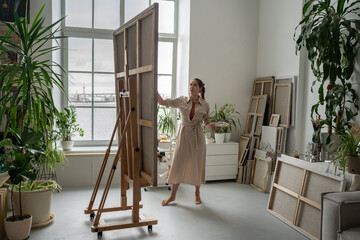 Inspire talented female artist in spacious home studio filled with green houseplants, canvas on floor and large windows painting picture on easel, modern art hobby therapy practice for mental health.