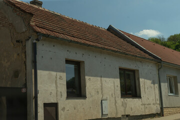 worn plaster house facade under sunlight showing cracked walls and tiled roof. two windows set in patched exterior, visible gutter, drainpipe,