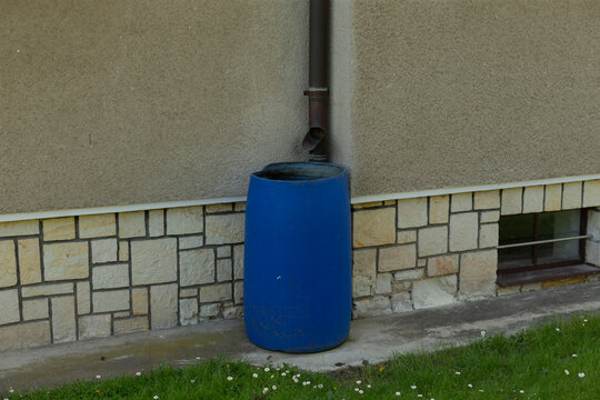 Blue rain barrel collecting downspout water beside stucco foundation and stone veneer, rusted metal downpipe directs runoff into plastic barrel, small