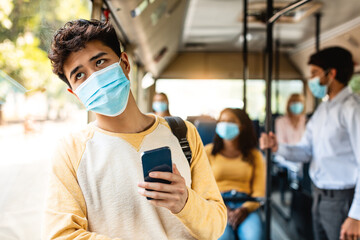 Public Transportation. Portrait of young asian man in protective medical mask using smartphone standing in city bus leaning on window. Bored male passenger looking out on street through window