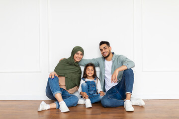 Portrait of cheerful arabian man, woman in headscarf and cute girl sitting on the floor leaning on white wall posing in new apartment in empty living room, family looking at camera, free copy space