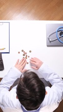 Vertical video doctor hands counting scattered coins on a white desk next to a stethoscope, symbolizing the financial burden of healthcare, costs, and medical debt