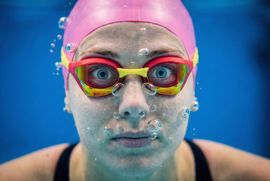 Funny underwater close-up of female swimmer face with wide eyes and bubbles wearing pink cap and goggles in blue swimming pool, representing surprise and immersion - Powered by Adobe