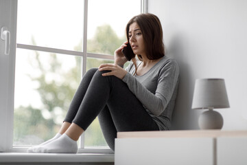 Sad upset depressed european millennial woman sitting on windowsill talking on phone, getting bad...