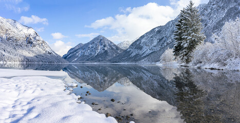 Atmospheric panoramic winter view of Heiterwanger Lake in Tyrol, Austria, surrounded by snowy alpine mountains and quiet winter nature. Calm and scenic winter mood in the European Alps.