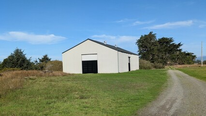 Obraz premium Modern white shed with open rollup doors sits along a hiking trail, surrounded by wind-shaped trees, grass, and blue skies, suggesting shelter, purpose, and quiet utility.