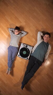 Vertical video father and son lying on a wooden floor together, enjoying a moment of connection and shared passion while listening to a vintage record player