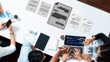A dynamic office meeting scene showing professionals engaged in discussion while analyzing data on laptops and documents scattered across the table. Raster