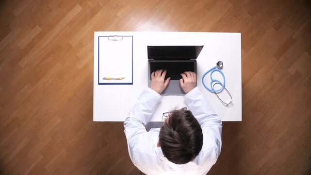 Doctor in lab coat opens laptop on white desk beside stethoscope and clipboard, preparing for a telemedicine consultation or virtual appointment in clinical office setting