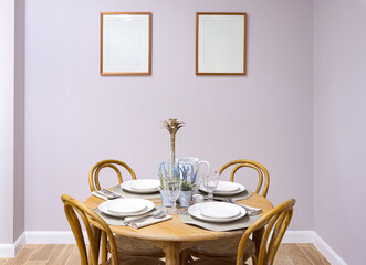 Dining room interior with wooden table set for four, chairs, and two blank frames on a light purple wall