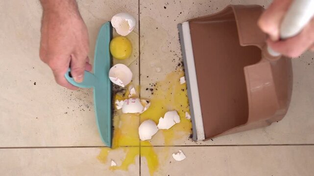 Cleaning broken eggs from a dirty tiled kitchen floor with a scraper and dustpan