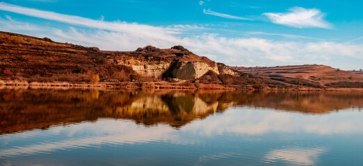 The scene captures the tranquil water beautifully mirroring the blue sky and white clouds above, as well as the steep, layered cliffs and hillsides of the local terrain