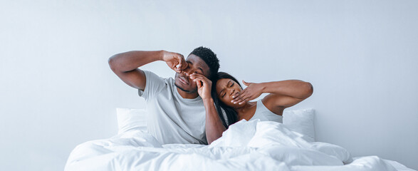 A couple is lying in bed, stretching and yawning as the morning light fills the bright room. They...
