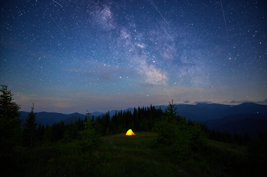 Single glowing tent on grassy hillside under stunning, star-filled sky with Milky Way and multiple shooting stars. Surrounded by pine trees and distant mountains. Beauty of clear night in nature. - Powered by Adobe