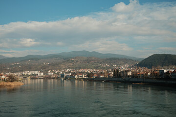 Fototapeta premium Wide view of the Drina River flowing past Visegrad with hills and residential buildings along the riverbank. Calm water and open sky emphasize the natural rhythm of the town.