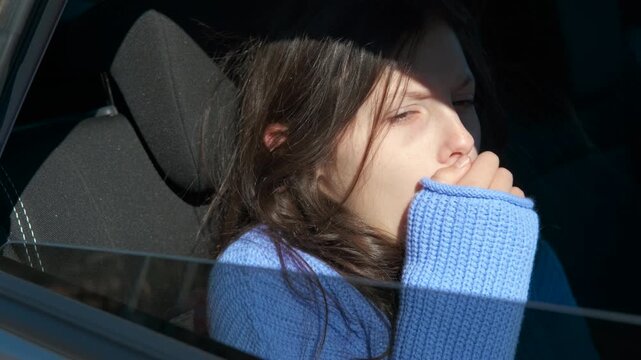 Sleepy young girl yawning in car during trip. Tired young girl in a blue sweater yawning while sitting in the passenger seat of a moving car, struggling to stay awake on a sunny day during a road trip