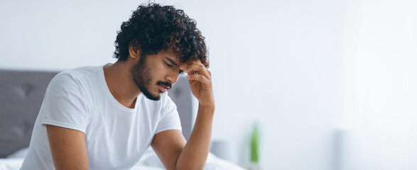 A young man sits on a bed with his hand on his forehead in a bedroom. The room is bright and airy,...
