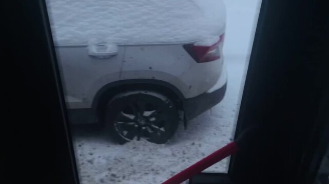bus interior snowy street view shows passing car gliding by, red safety handrail in foreground, snowcovered curb and slushy pavement, muted Taken on mobile device.