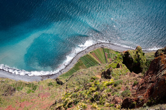 Top down view from the Cabo Gir&atilde;o skywalk on Madeira Island, Portugal, shows terraced fields on a narrow coastal strip far below steep red cliffs with turquoise Atlantic Ocean waves.