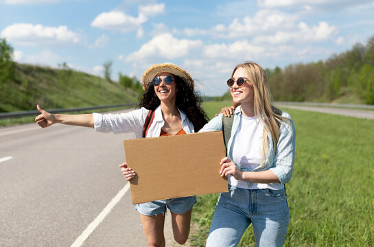 Two female friends hitching free ride, holding blank carton sign with mockup, stopping car on road, free space. Lovely young women enjoying summer freedom, traveling by autostop