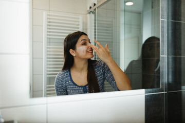 Beautiful young woman applying moisturizing face cream in front of a bathroom mirror