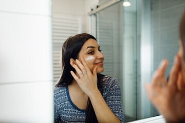 Beautiful young woman applying moisturizing face cream in front of a bathroom mirror