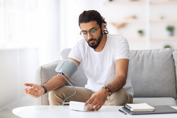 Young arab man sitting on couch, checking blood pressure and taking notes at home. Indian guy in...