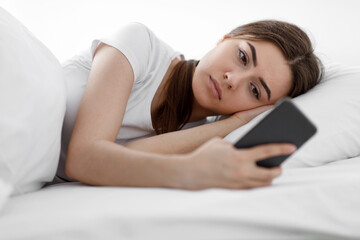 Fototapeta premium Sad sorrowful caucasian millennial lady, reading message on smartphone, waiting for sms on bed in bedroom at home, copy space. Chat online, gadget addiction, depression, insomnia and sleep problems
