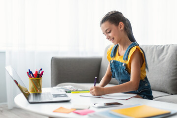Portrait of smiling schoolgirl sitting on couch at desk with pc, writing essay or drawing picture in paper notebook. Pupil studying or taking notes in her diary, making checklist, free copy space