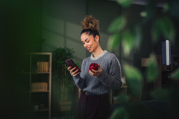Woman smiling while multitasking, enjoying healthy snack at home