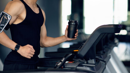 Naklejka premium Sporty Lifestyle. Unrecognizable Male Athlete Jogging On Treadmill At Gym, Cropped Shot Of Young Man Holding Fitness Shaker Bottle Training On Modern Equipment At Sport Club, Panorama With Free Space