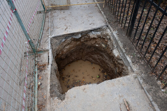 Deep hole excavated in a concrete sidewalk, filled with muddy water and fallen leaves, next to a temporary construction fence