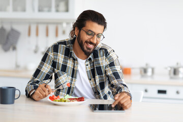 Positive long-haired, bearded arab guy having breakfast, eating fresh vegetables, sausages, drinking coffee and using modern digital tablet, watching series or videos online, kitchen interior