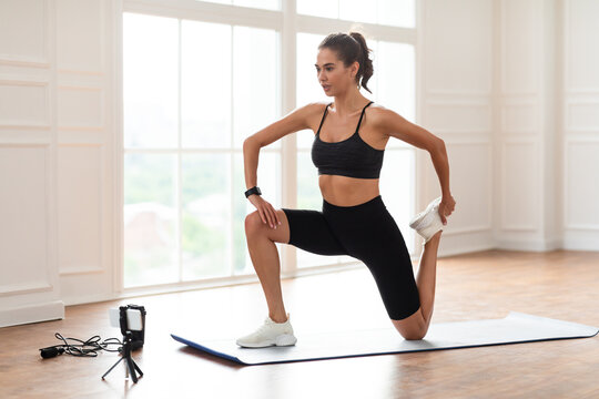 Healthy Sporty Lifestyle. Focused fit lady stretching back leg muscles and quads holding feet watching online tutorial or recording fitness video for her sport blog, using cell phone on tripod