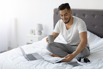 Young Arab freelancer sits in bed with a laptop and notebook, checking his schedule while enjoying coffee. He embraces remote work opportunities in a comfortable setting.