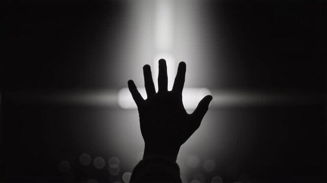Silhouette of a hand raised in worship towards a glowing cross. Black and white footage of a person praying in a dark church. Christian faith and spirituality concept