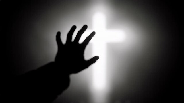 Silhouette of a hand reaching towards a glowing cross. Black and white spiritual footage of faith and prayer. Religious concept with bokeh candle lights