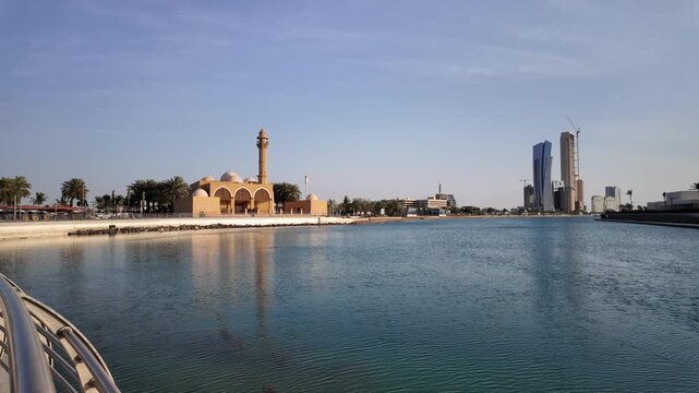Serene waterfront mosque reflecting on calm waters of Jeddah Corniche, with modern skyscrapers rising in the distance under a clear blue sky at sunset