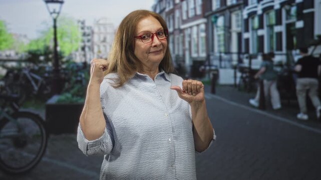 Woman with red glasses points thumb to chest and flexes fist on a street beside bicycles and row houses under a lamppost wearing blue checkered shirt; confidence.