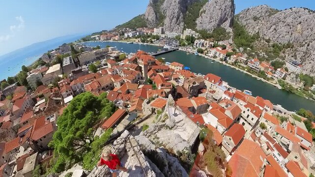 Woman stands on Mirabella Fortress, Peovica, overlooking Omis with red-tiled roofs, the Cetina River meeting the Adriatic, cliffs and mountains under a clear blue sky, summer panorama in Croatia