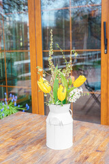 Bouquet of fresh spring flowers including yellow tulips arranged in white metal vase on wooden table in outdoor cafe with blooming trees. Warm light, garden reflections, and seasonal floral decoration