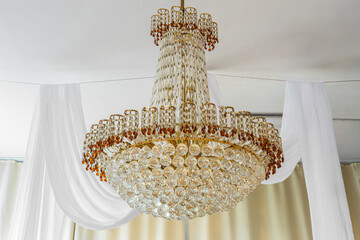 Beautiful luxury crystal chandelier with golden details hanging from the ceiling reflecting light and creating luxurious interior atmosphere in room with white curtains